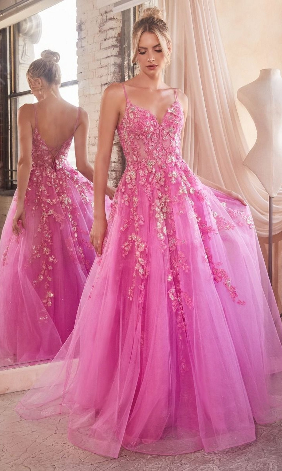 A woman stands in front of a mirror in a softly lit room, wearing the Long Prom Ball Gown with Flowers CM347—a floor-length pink dress embellished with floral details and featuring spaghetti straps.